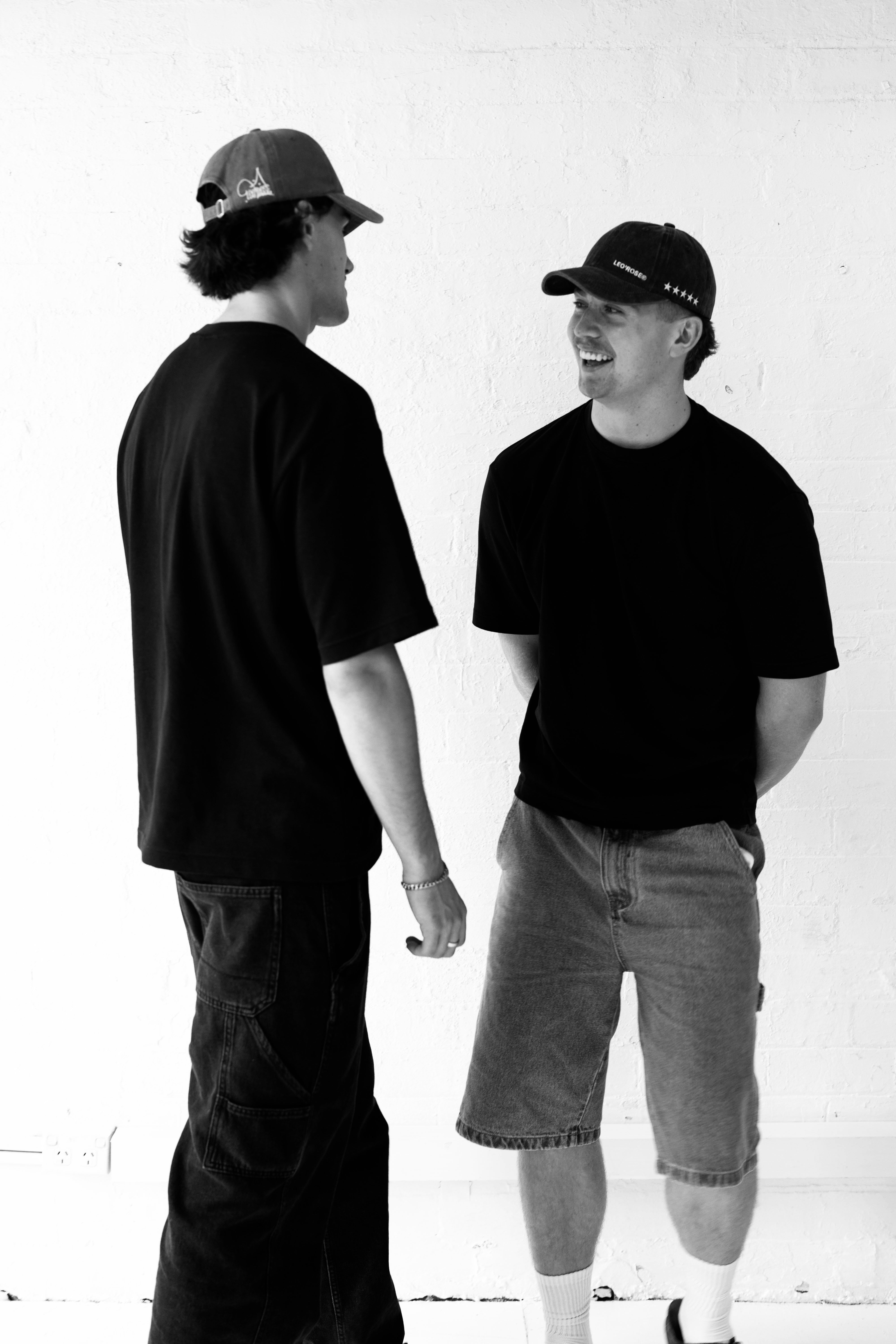 Two males, both wearing LEO'ROSE hats, looking at each other with a smiling expression